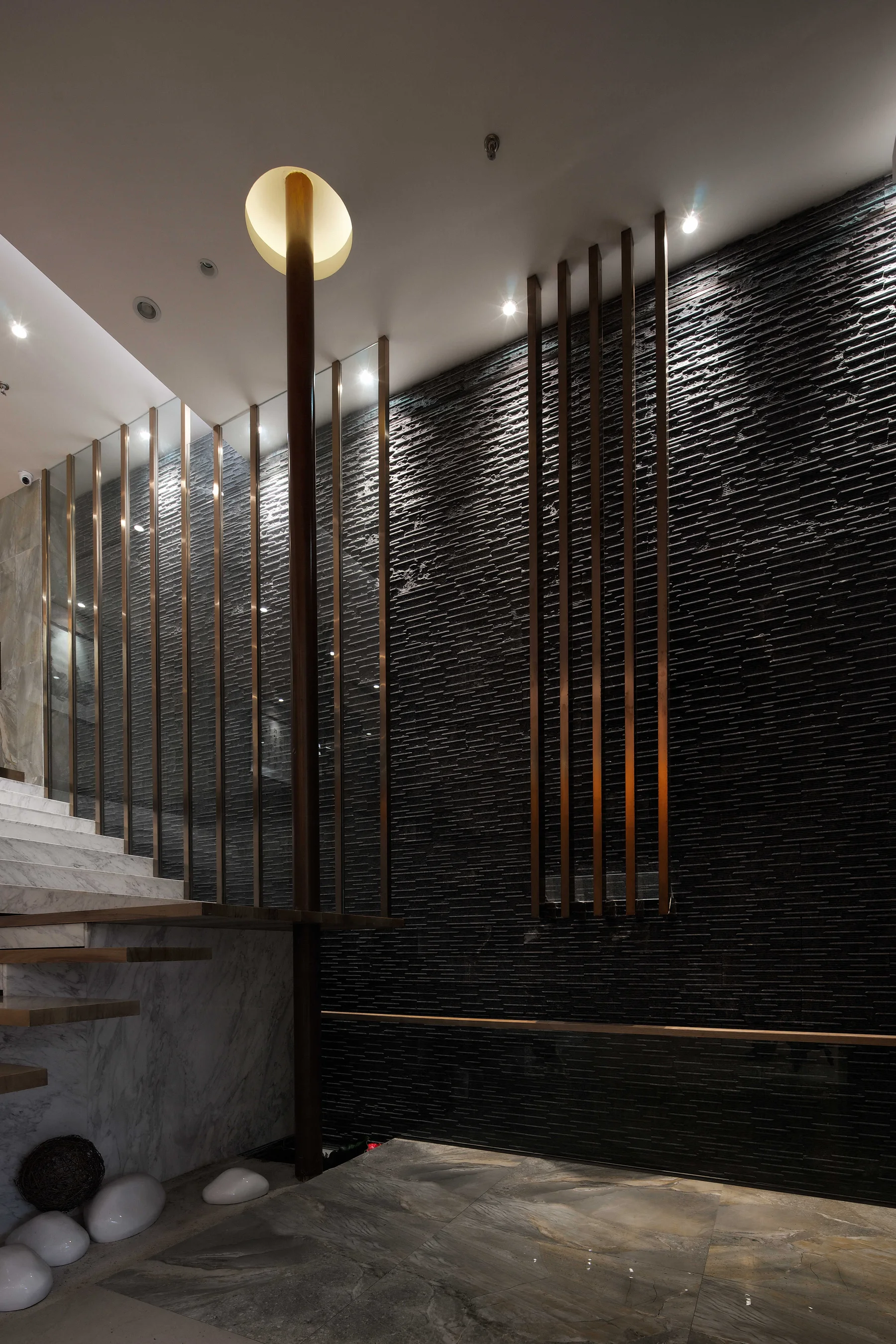 Textured black stone wall crossed by bronze fins and a circular light